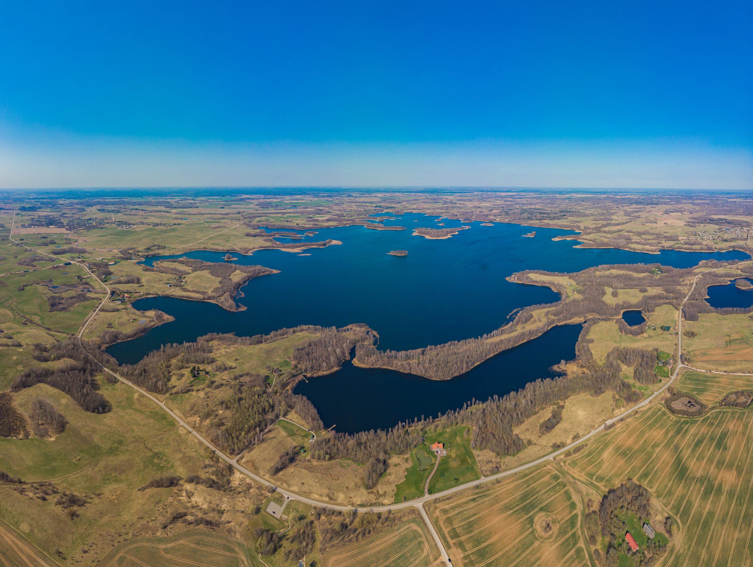 Aerial view of Lake Rubikiai in spring, its deep blue water scattered with wooded islands among fields and forest